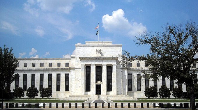 The Federal Reserve Building in Washington D.C., showing its iconic white marble neoclassical architecture with columns, an eagle statue, and the American flag flying on top.
