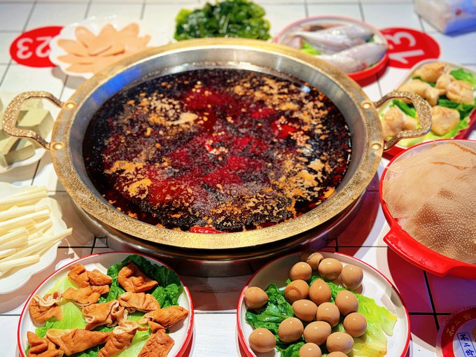 A spread of Chinese hot pot ingredients including a large pot of red spicy broth, plates of quail eggs, meat, tofu, vegetables, and dumplings on a white tiled table.