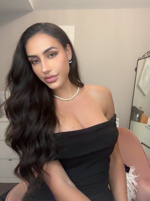 A woman with long, dark wavy hair poses in a black off-shoulder dress and pearl necklace.
