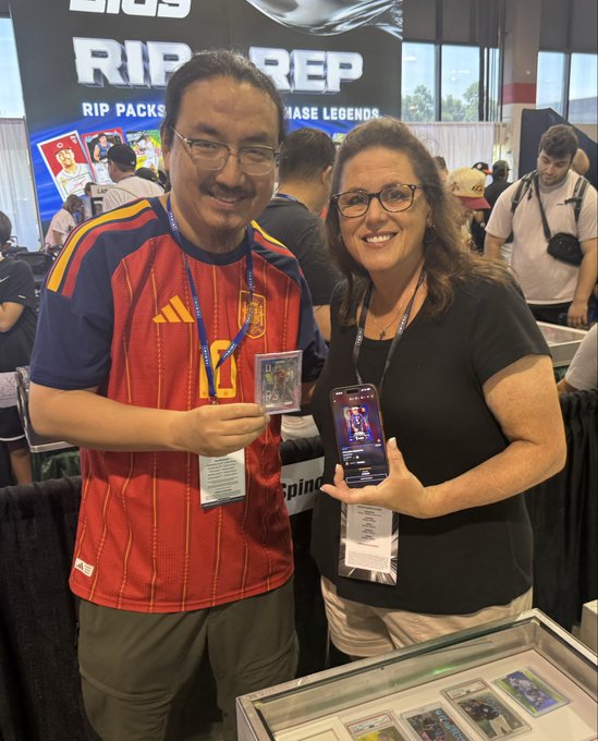 Two people smiling and posing at a 'Topps RIP REP' trading card convention booth, holding up a sports card and showing it on a smartphone.