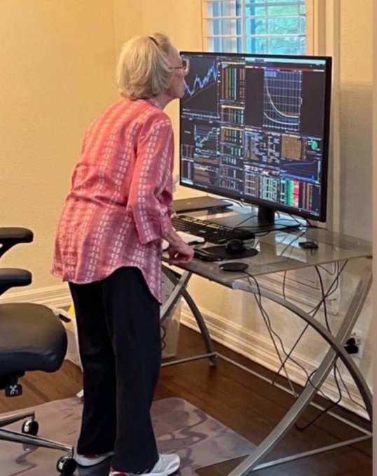 An elderly woman standing at a desk examining a large monitor displaying multiple trading charts and graphs.