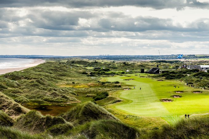 A scenic view of a coastal golf course with rolling sand dunes, a green fairway, and a beach under a cloudy sky.