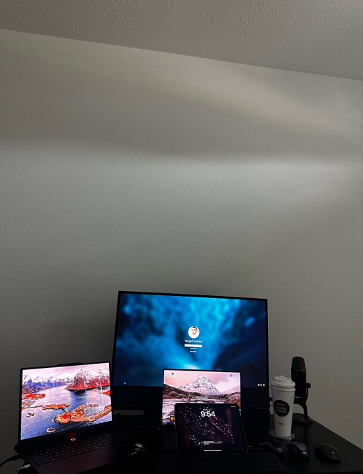 A dimly lit home office setup featuring a laptop, monitor, iPad, and a coffee cup, all displaying matching mountain landscape wallpapers.