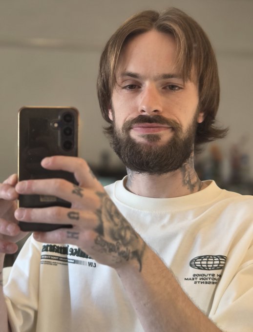 A bearded man with shoulder-length brown hair takes a mirror selfie with a black Samsung smartphone, showing off tattoos on his hands and wrist. His white "Lacoste" t-shirt and steadily growing locks visually document his commitment to the haircare challenge tied to XRP's price milestone.