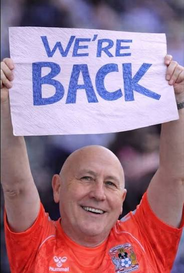 A bald man in an orange Huddersfield Town football shirt smiles broadly while holding up a handmade "WE'RE BACK" sign, visually echoing the post's celebration of Bitcoin reclaiming the $64K level.