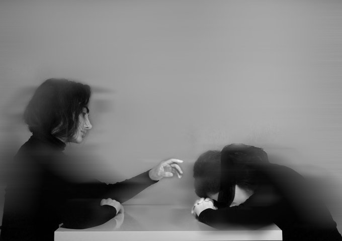 A black and white image depicts two figures in motion blur; one reaches an outstretched hand toward another person hunched over a table with their head down.