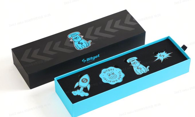 A Bitget promotional gift box set featuring a black outer sleeve with a mascot graphic and a turquoise inner tray containing branded enamel pins.