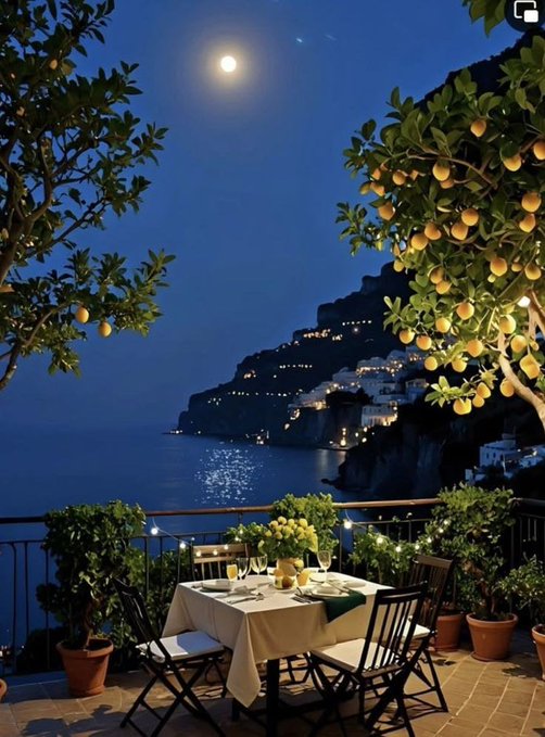 A scenic evening view from a balcony in what appears to be a Mediterranean coastal town, likely the Amalfi Coast, features a table set for two with wine glasses and lemon trees framing the moonlit sea.
