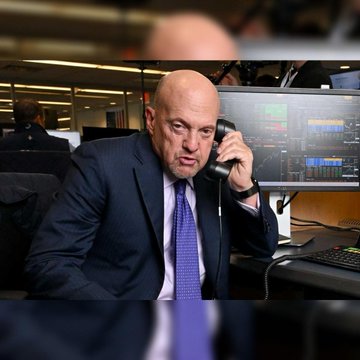 A bald stock trader in a dark suit and blue tie speaks on a phone amid a backdrop of multiple financial trading screens displaying market data, visually capturing the Wall Street trading floor environment referenced in the post about NYSE and Nasdaq moving toward extended 23×5 trading hours to meet growing demand beyond traditional sessions.