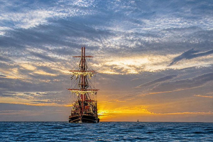 A tall sailing ship is silhouetted against a vibrant sunset over the ocean, with dramatic clouds filling the sky.