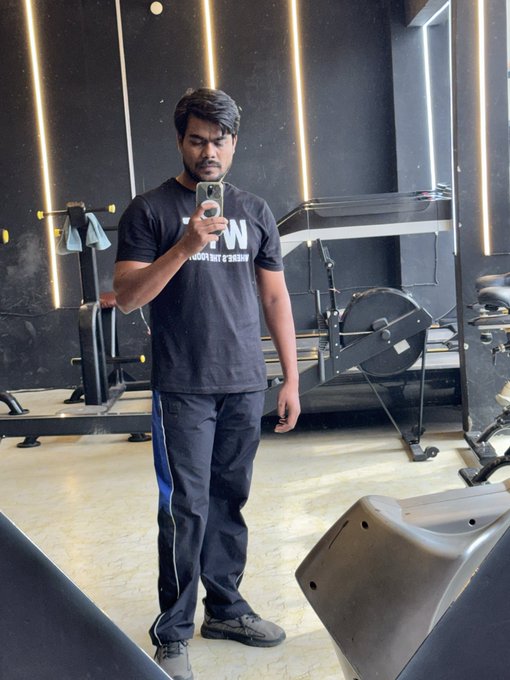 A man takes a mirror selfie in a gym, wearing a black "FROST" t-shirt and dark track pants, with workout equipment visible in the background.