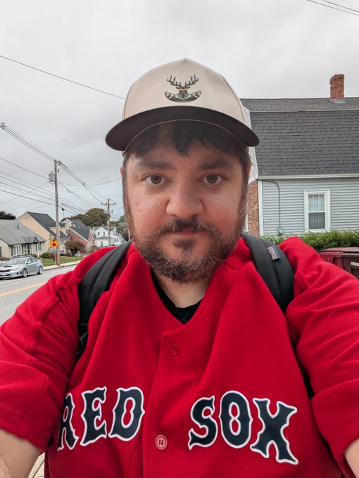 A selfie of a bearded man wearing a tan trucker hat with a deer logo and a red baseball jersey with 'RED SOX' written across the chest, standing on a residential street with houses in the background.