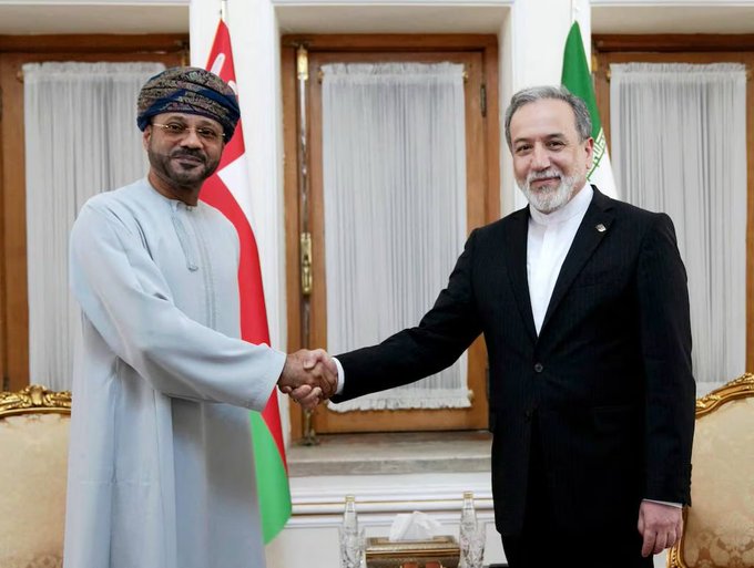 Two men shake hands in a formal setting; the man on the left wears traditional Omani attire with a patterned turban, while the man on the right wears a dark suit, with Omani and Iranian flags visible in the background.