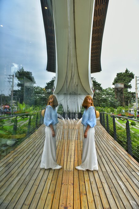 A mirrored photograph of a woman in a white dress and blue top standing on a wooden boardwalk in front of a modern architectural structure with a tall central pillar.