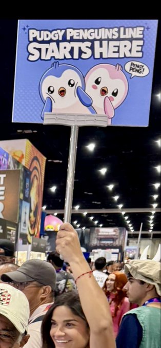 A person holds up a sign at a convention reading 'Pudgy Penguins Line Starts Here' featuring two cartoon penguin characters, one calling out 'PENGU PONG!'.
