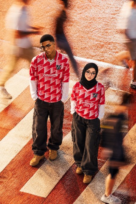 Two people wearing matching red and white Budweiser-style patterned jerseys and dark cargo pants stand on a red crosswalk, with blurred figures of other people moving around them.