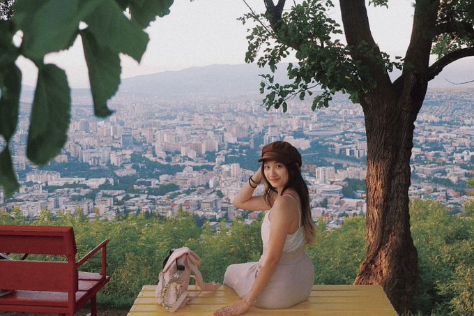 A young woman in a white dress and brown cap sits at a scenic overlook above a sprawling city, with a red bench and tree framing the view. The image evokes a peaceful, reflective moment that aligns with her warm, encouraging message to her audience, suggesting a personal break to recharge and connect with others.
