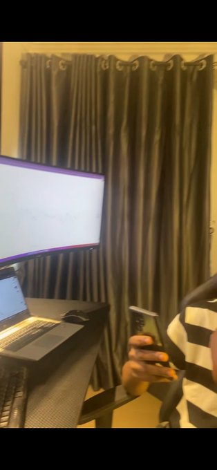 A dimly lit bedroom selfie showing a person with long yellow nails holding a smartphone, with a curved monitor displaying a light screen, a laptop showing a chart with '(+3.24%)' visible on the left, and dark curtains in the background.