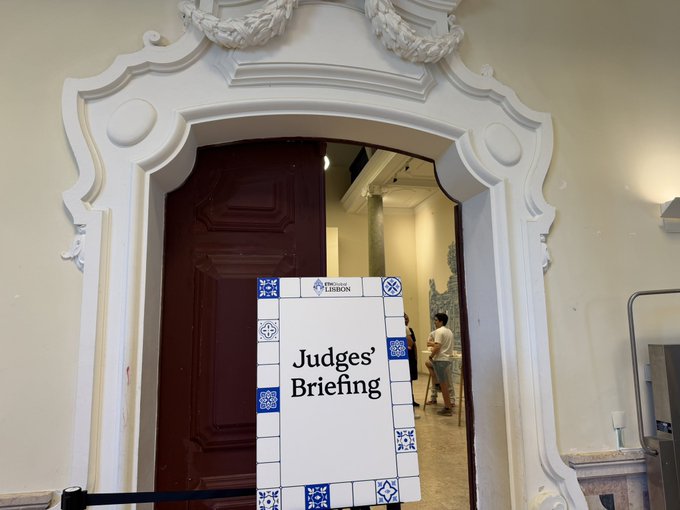 A decorative white ornate archway frames a doorway with a "Judges' Briefing" sign featuring ETHGlobal Lisbon branding, where attendees can be seen gathering in the room beyond for presentations evaluating the overnight builders' projects. The elegant baroque-style doorway with its dark wooden door sets a formal tone for the judging sessions at this Ethereum hackathon.