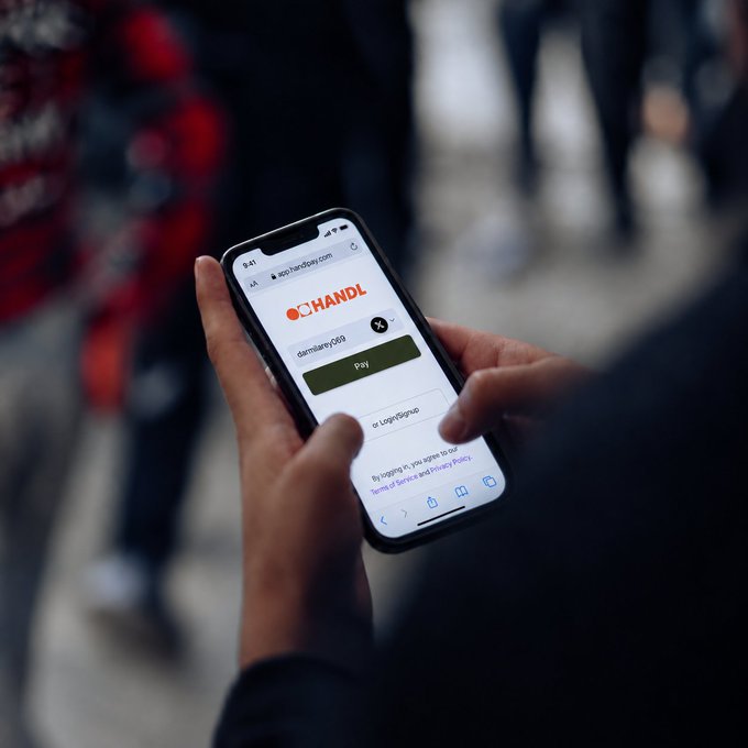 A person uses a smartphone displaying a payment app called 'HANDL' with the URL 'app.handlapp.com' to send 26.5 dollars to a 'Logan Group' account in a 'Beneficiary pass.'