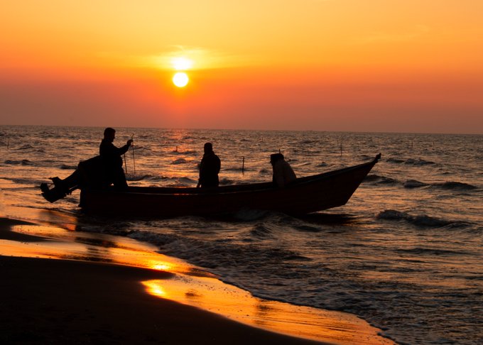 A silhouetted scene of three fishermen in a small boat on the water at sunset, with the orange sky reflecting on the waves and beach.