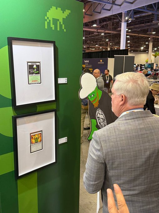 An older man in a gray plaid suit viewing framed Magic: The Gathering cards displayed at a convention booth, with a Space Invaders-style alien logo on the green wall above.