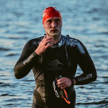 A bearded man wearing a red swim cap and black wetsuit emerging from the water, adjusting his goggles after an open-water swim.