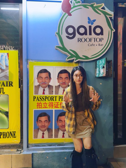 A woman in a yellow plaid shirt and shorts poses next to a 'gaia ROOFTOP Cafe Bar' sign and a 'PASSPORT PHOTO' booth featuring Mr. Bean's face.