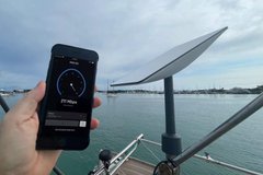A hand holds a smartphone displaying a "Pi" branded app interface with a speedometer-like gauge, set against a backdrop of a satellite dish and water under a cloudy sky—visually linking the Pi Quantum Network blockchain project to themes of global connectivity and advanced technology.