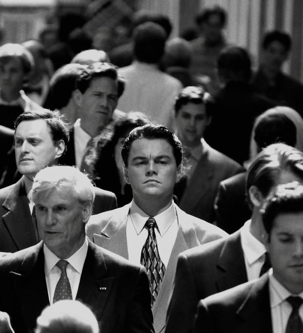 A black-and-white photograph showing a crowded scene with a young Leonardo DiCaprio in a suit and patterned tie standing out among a group of businessmen in formal attire.