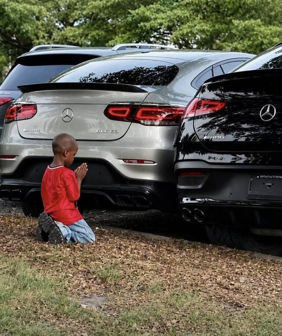 A young child in a red shirt and blue jeans kneels on grass and dirt in front of two parked Mercedes-AMG cars, with their hands clasped in prayer.