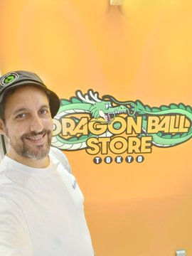 A smiling bearded man wearing a dark cap taking a selfie in front of an orange wall with the 'Dragon Ball Store Tokyo' logo.