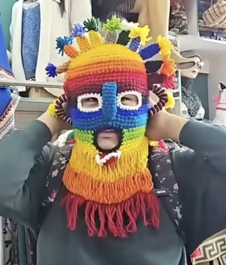 A person wearing a colorful crocheted balaclava mask featuring rainbow stripes and dreadlock-like fringes in red, yellow, blue, and green colors, displayed in what appears to be a clothing store.