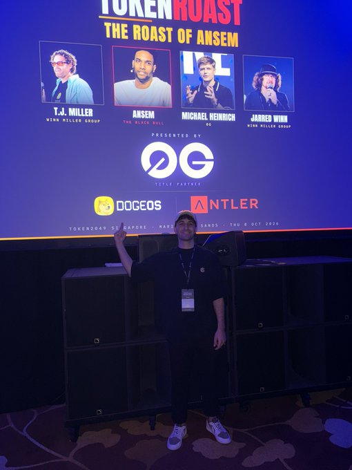 A young man wearing a black cap, lanyard, and white sneakers points toward a large event screen for "TOKEN ROAST: THE ROAST OF ANSEM," featuring comedians T.J. Miller, Ansem (The Black Bill), Michael Kenneihrich, and Jared Winn, presented by OG with title partners DogeOS and Antler. The image relates to the post, as the man appears excited to attend the event roasting Ansem, a controversial crypto personality known for his Solana price predictions.