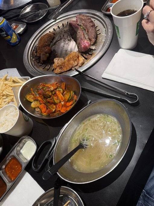 A Korean BBQ meal spread featuring grilled meat on a tabletop grill, vegetable side dishes, fries, rice, and a bowl of noodle soup.
