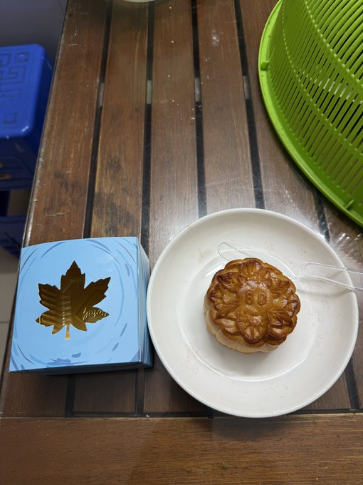 A mooncake sits on a white plate next to a blue gift box with a leaf design on a wooden slatted table, alongside a green colander and blue crate.