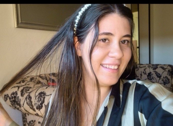 A smiling selfie of a woman with long dark hair wearing a pearl headband and a black and white striped shirt, sitting on a patterned couch.