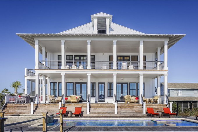 A large, two-story white house with a wrap-around porch, multiple Adirondack chairs, and a swimming pool sits under a clear blue sky.