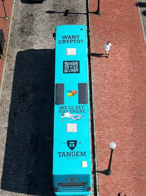 An aerial view of a teal Tangem-branded bus on a brick street with advertising reading 'WANT CRYPTO? WE'LL GET YOU THERE' and a QR code on top, along with 'TANGEM TRUST & SECURITY 2026' branding. A person walks on the sidewalk nearby.