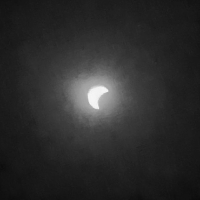 A black and white photograph of a partial solar eclipse, showing the sun crescent visible through thin clouds in the night sky.