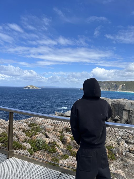 A person in a black hoodie stands with their back to the camera, gazing out at a scenic ocean view with rocky cliffs and blue sky.