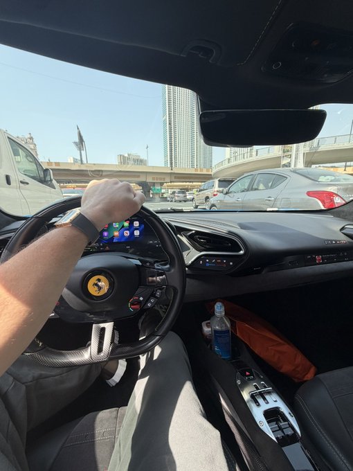 A driver grips the steering wheel of a Ferrari, wearing an Apple Watch, as they navigate through traffic on a busy multi-lane highway. The luxury sports car interior and confident driving posture contrast with the post's message about taking bold, unconventional action in a rigged crypto market.