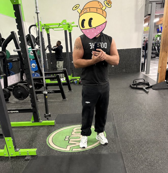 A man with his face covered by a cartoon emoji sticker stands in a gym wearing a black tank top that reads 'EQRE' and black sweatpants.