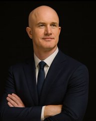 A bald man in a navy blue suit and black tie posing with crossed arms against a dark background.