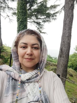 A selfie of a woman wearing a patterned headscarf, standing outdoors among tall evergreen trees.