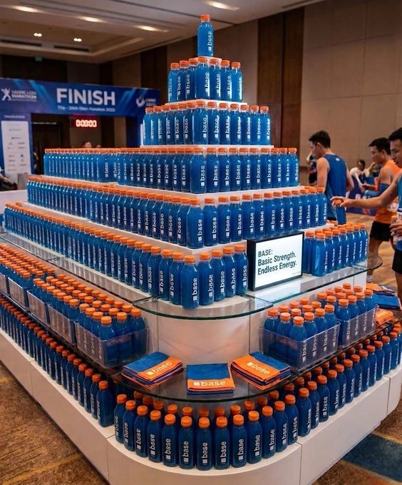 A large pyramid-shaped promotional display features hundreds of blue "BASE" electrolyte bottles with orange caps, with a single bottle crowning the top tier, while a small sign reads "BASE: Basic Strength. Endless Energy." The setup appears at a marathon finish line area, fitting the "Baseposting" theme of showcasing the Base hydration brand's athletic sponsorship.