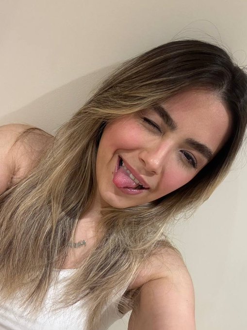 A young woman with long, wavy highlighted hair winks playfully at the camera and sticks her tongue out.