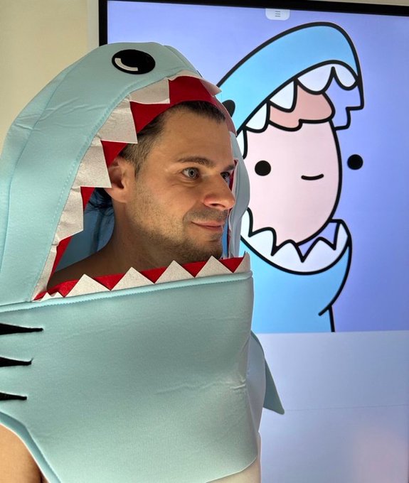 A man smiles while wearing a light blue shark costume, with a cartoon image of a similar shark displayed on a screen behind him.