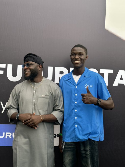 Two smiling men pose at an event, including a tall man in a bright blue shirt giving a thumbs-up beside another man in a gray traditional outfit and sunglasses.
