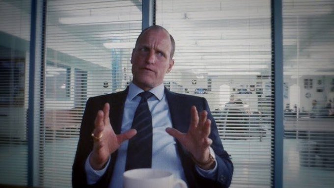 A man in a suit sits in an office with venetian blinds behind him, gesturing with both hands raised while speaking.